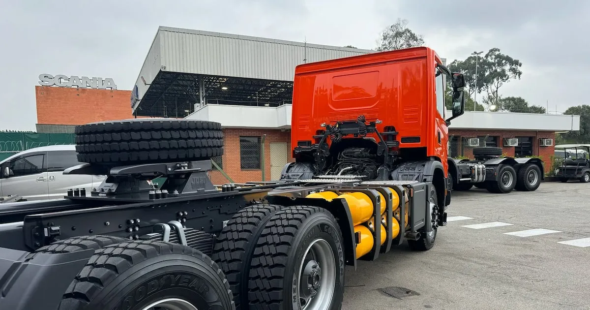 Detalhe da banda de rodagem de um pneu de caminhão com veículos de carga ao fundo em uma rodovia